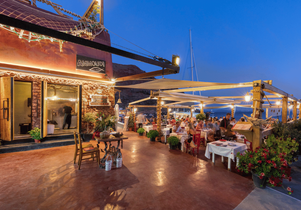 A vibrant Santorini restaurant at night, with patrons dining outdoors under warm lights in Ammoudi, Oia