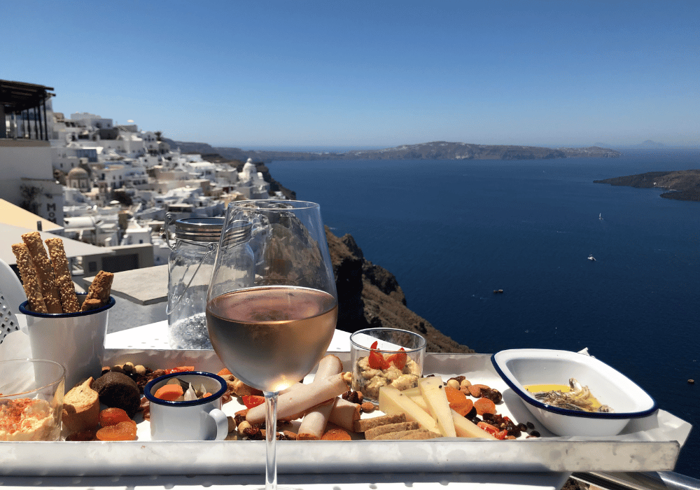 A beautifully arranged tray with food and wine on a table, offering a stunning view of the sea in Santorini
