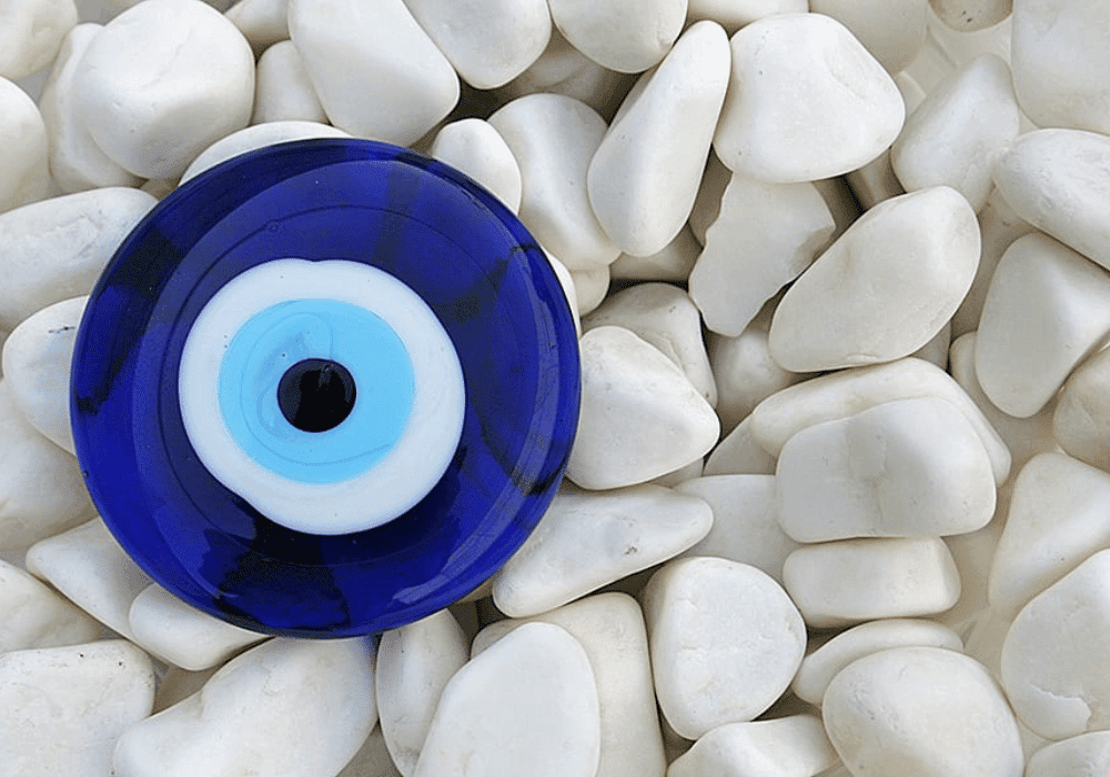 A blue evil eye ornament is positioned on white pebbles, embodying protection and positivity at Erossea Villa Santorini Shopping