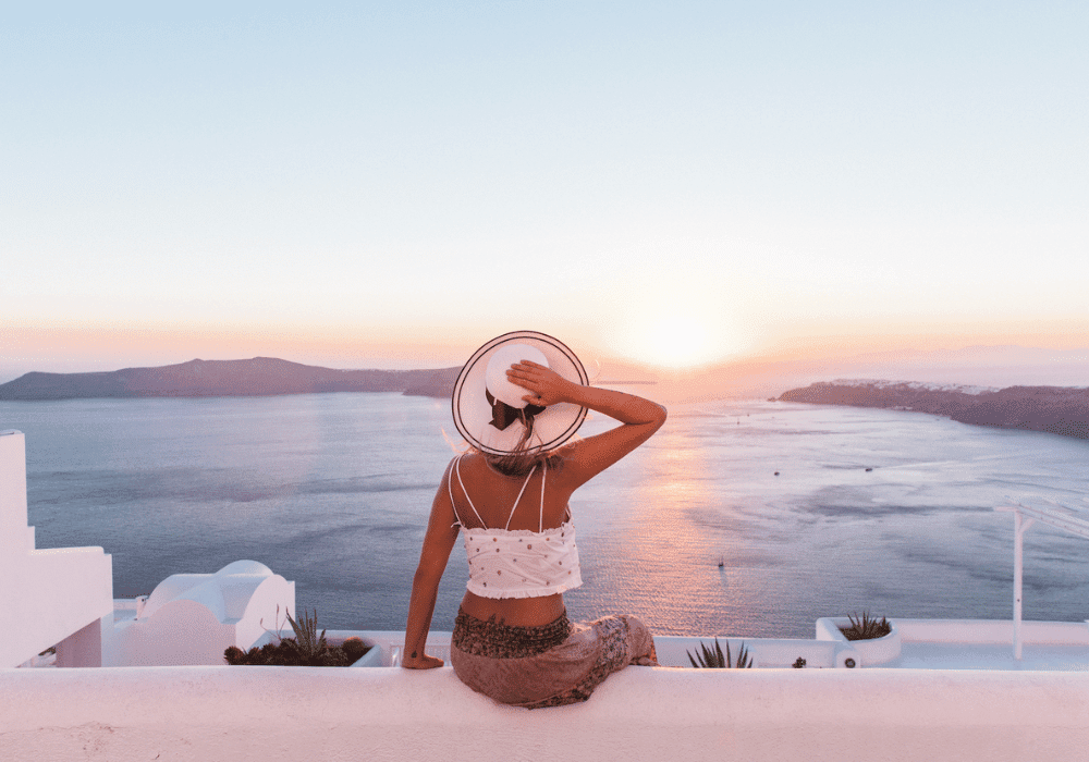 A woman in a hat and summer clothes sits on a ledge, gazing at the ocean from Erossea Villa Santorini