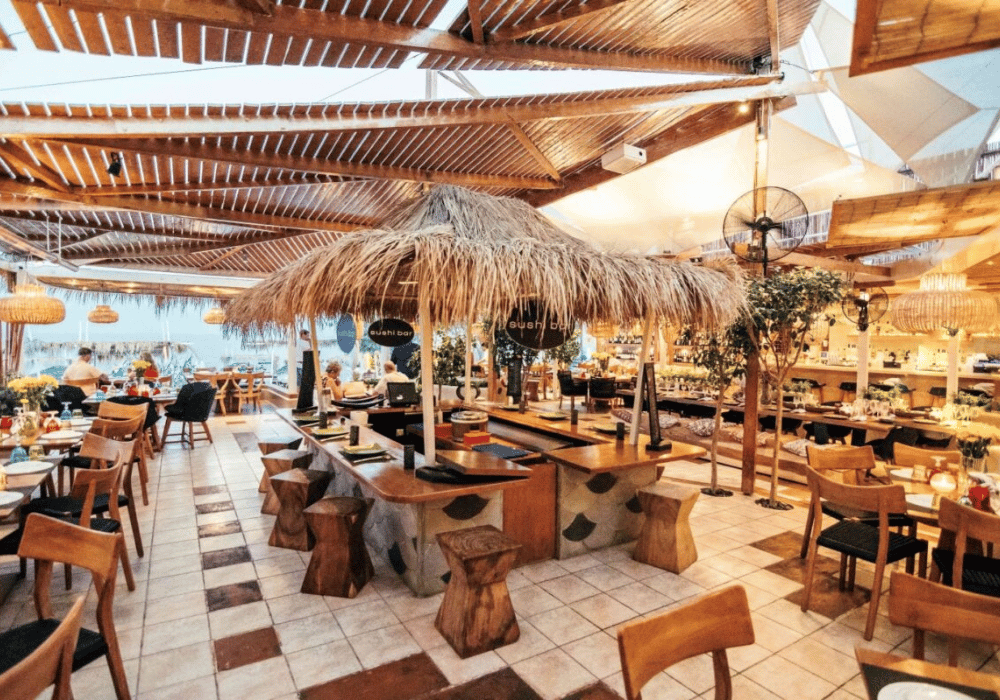 A seaside restaurant in Santorini featuring wooden tables and a thatched roof, offering Mediterranean cuisine with a modern flair.